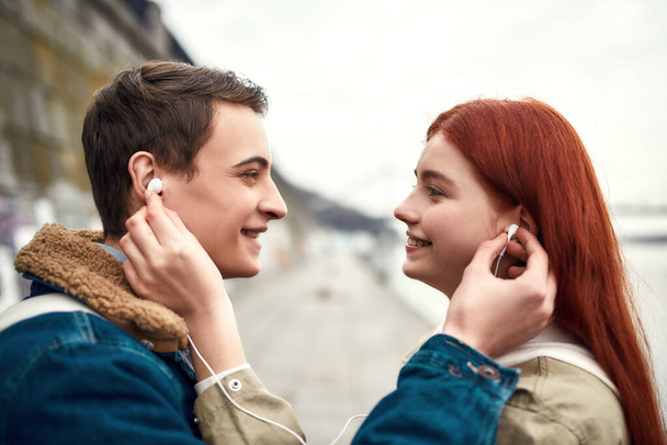 Close up of couple of teenagers going to listen to music using the same pair of earphones, looking at each other with love - Фото, изображение