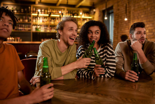 Friends in the bar watching sports match on TV together, drinking beer, clinking bottles and cheering for team. People, leisure, friendship and entertainment concept - Photo, image