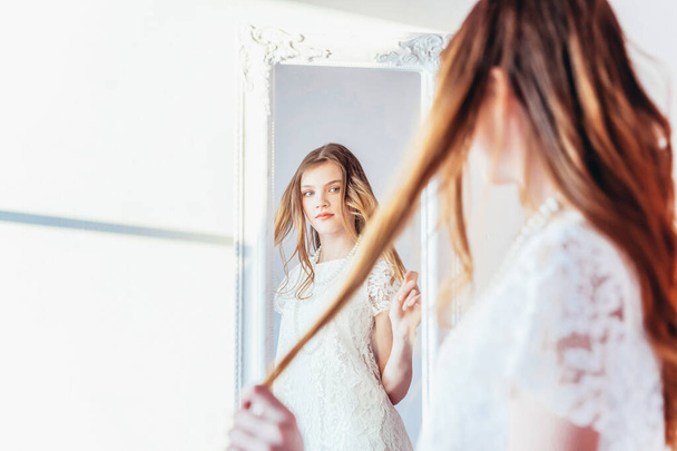 Красота макияжа утром - любить себя. Молодая девушка-подросток смотрит на отражение в зеркале. Молодая позитивная женщина в белом платье позирует в ярком светлом помещении напротив белой стены - Фото, изображение