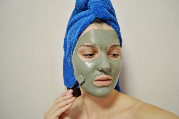 Μια γυναίκα με βούρτσα βάζει μια πήλινη μάσκα στο πρόσωπό της με μπλε πετσέτα στο κεφάλι της. - Φωτογραφία, εικόνα