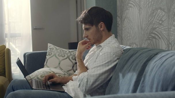 Vážný muž sedící na gauči s počítačem. Zaměřený chlap pracující na notebooku uvnitř - Fotografie, Obrázek