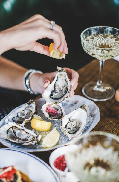 Hands of woman squeezing lemon juice to Irish oysters in plate with ice over glass of champagne in fish restaurant at background, selective focus. Seafood, French cuisine, fine dining concept - Foto, imagen