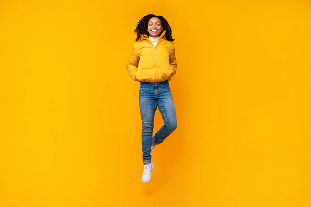 Joyful African Woman Jumping Wearing Winter Jacket Over Yellow Background - Fotografie, Obrázek