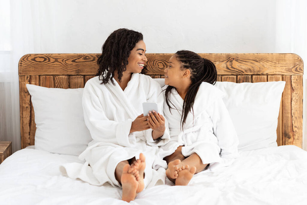 Afrikanische Mutter und Tochter mit Smartphone beim Entspannen auf dem Bett im Bademantel - Foto, Bild