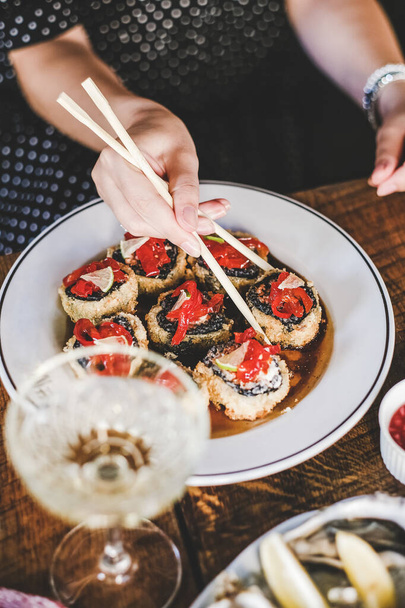 Girl eating baked susi maki with chopsticks over fresh oysters and champagne on table at Japanese restaurant, top view, selective focus. Japanese cuisine, fast food, seafood, fine dining concept - Photo, image