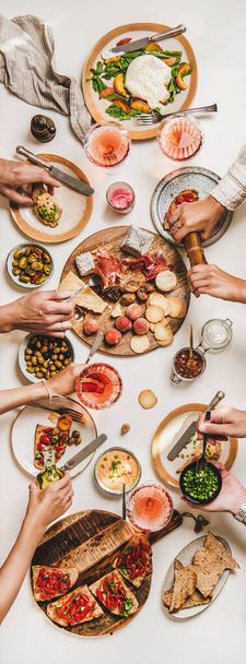 Family wine and snack party. Flay-lay of peoples hands eating and drinking rose wine over table with cheese, fruit, smoked meat, tomato brushettas, buratta salad, top view. Wine tasting concept - Zdjęcie, obraz