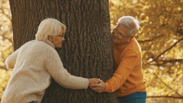 Το ηλικιωμένο ζευγάρι έχει ρομαντική φθινοπωρινή μέρα στο δάσος. Αγκαλιάζοντας δέντρο και χαμογελώντας - Φωτογραφία, εικόνα