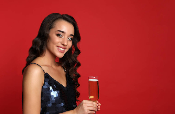 Beautiful young woman in elegant dress holding glass of champagne on red background. Christmas party - Foto, Imagen