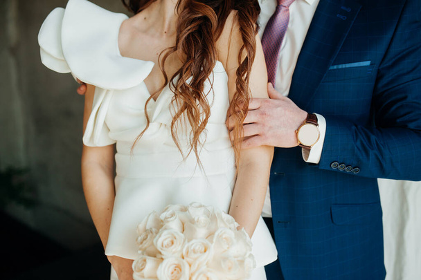 Κομψή φωτογραφία γάμου. Ο γαμπρός σε μπλε κοστούμι αγκαλιάζει απαλά τη νύφη σε ένα λευκό κομψό φόρεμα από σκληρό ύφασμα. Φουντωτό νυφικό μανίκι, flounce μανίκι σε ένα ασυνήθιστο νυφικό. - Φωτογραφία, εικόνα