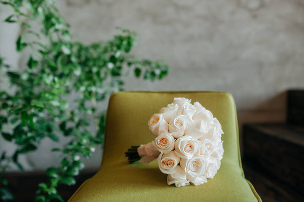 A stylish bouquet of white identical fresh roses on a light green chair against a gray concrete wall and a home green flower. - Фото, изображение