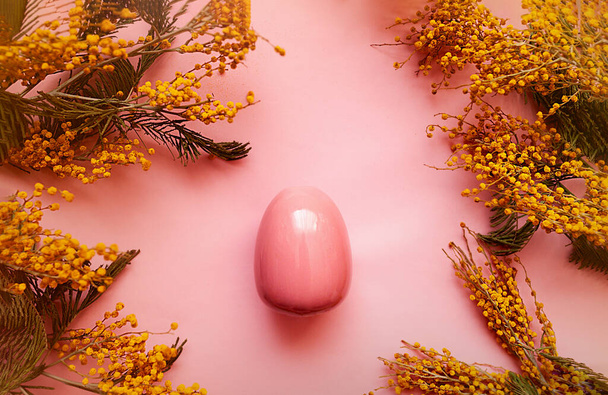 Frühlingskarte für einen frohen Osterurlaub. Rosafarbenes, glänzendes, sauberes Ei auf einem rosafarbenen Hintergrund in einem Rahmen frischer, sattgelber Mimosa-Blumen. - Foto, Bild