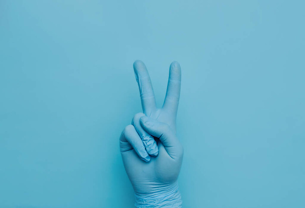 A hand in a blue medical surgical protective glove shows the figure peace with its fingers. Communication using gestures. - Foto, Bild