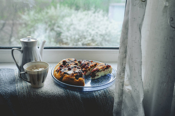 уютный дом чаепитие за окном в плохую погоду. Фарфоровый белый чайник и чашка чая, домашний ягодный пирог на круглой синей тарелке против цветущего дерева на улице за окном с занавеской - Фото, изображение