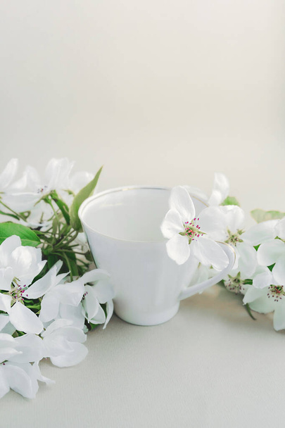 White porcelain cup with green tea and a fresh white pear tree flower in a lush bouquet of blooming white flowers and green bright leaves on a twig on a light table. vertical image - Фото, изображение