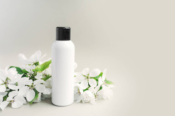 white clean bottle with a black cap with a blossoming white sprig of lush flowers with green leaves on white background. Cosmetic layout. Skin and body care mock up - Photo, Image