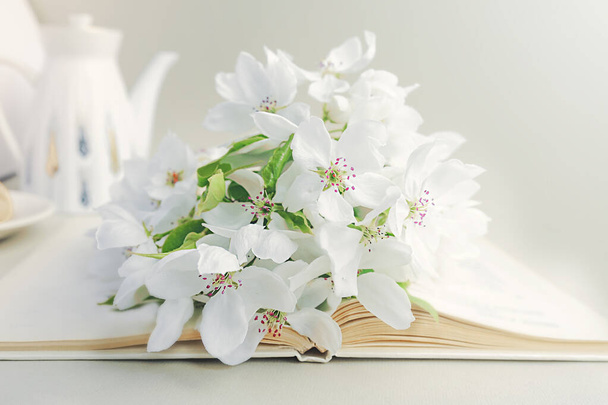 lush white bouquet of fresh flowers with green leaves on an open white paper, white porcelain teapot on the background. Reading time, summer bright morning - Photo, Image