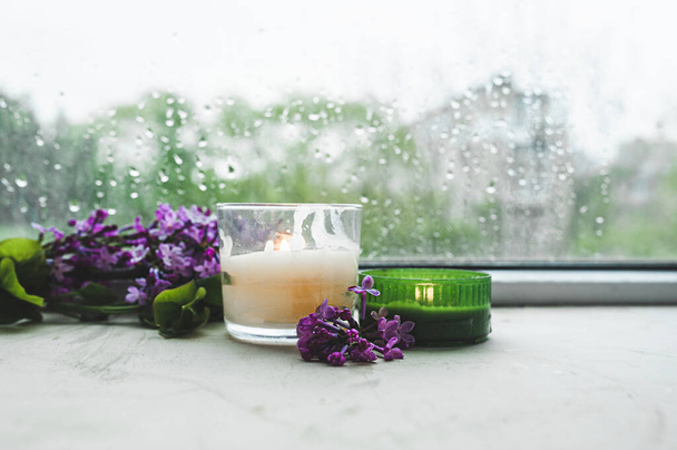 queimando duas velas branco e verde com ramos de flor lilás roxo fresco raminho no fundo de vidro molhado em gotas e gotas da janela - Foto, Imagem