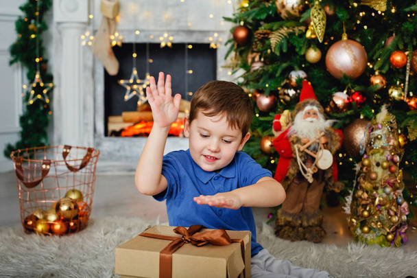 Cute boy at the Christmas tree and fireplace, decorated with garlands and gifts. New year mood. Bokeh of lights. - Photo, image