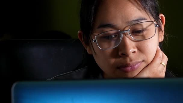Κουρασμένη γυναίκα φορώντας γυαλιά χρησιμοποιώντας την τεχνολογία φορητού υπολογιστή, ενώ κάθεται στο τραπέζι τη νύχτα. Νεαρή γυναίκα χασμουριέται νυσταγμένη λόγω σκληρής δουλειάς υπερωρίες στο σπίτι. εργασία στο σπίτι έννοια. - Πλάνα, βίντεο