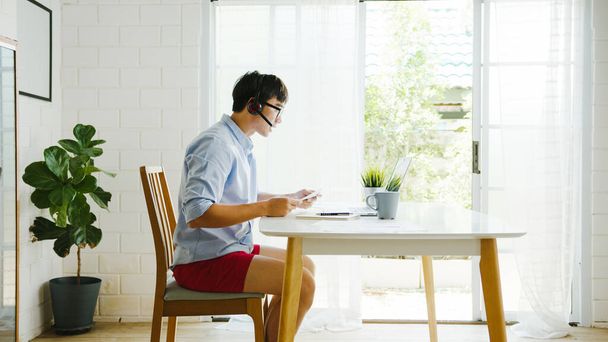 Asia businessman dressed in shirt and shorts use laptop talk to colleagues in video call while work from home at living room. Self-isolation, social distancing, quarantine for corona virus prevention. - Foto, imagen