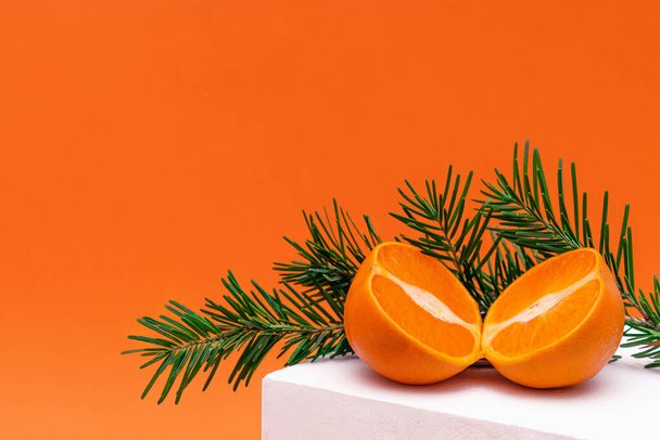 Modern still life with tangerine cut in half and fir branches on white podium. New Year,Christmas and winter concept with food and geometric objects on orange background.Copy space for text - Zdjęcie, obraz
