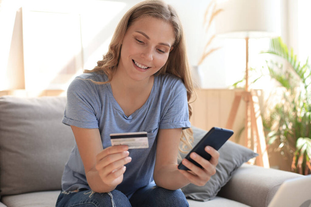 Happy natural blonde using credit card and mobile phone in living room - 写真・画像