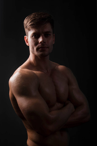 bodybuilder musculaire posant avec un torse nu sur un fond noir dans l'obscurité avec rétro-éclairage. Homme sexy. - Photo, image