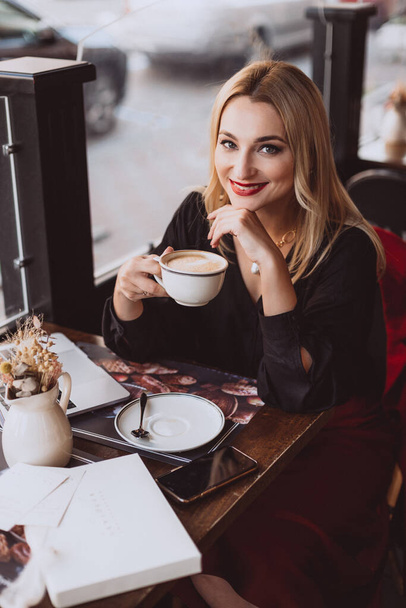 Piękna młoda kobieta biznesu z laptopem pije filiżankę kawy w kawiarni podczas jesiennej przerwy obiadowej. Miękkie wybiórcze skupienie, artystyczny hałas. - Zdjęcie, obraz