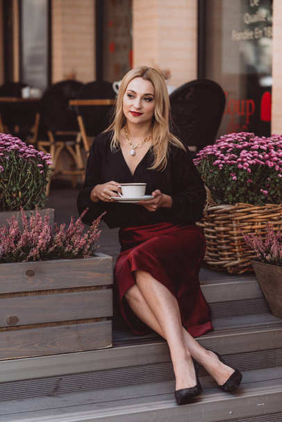 Beautiful young blonde woman in a trendy autumn outfit in burgundy marsala color with a cup of coffee on the steps of a cafe. Soft selective focus. Artistic noise. - Photo, Image