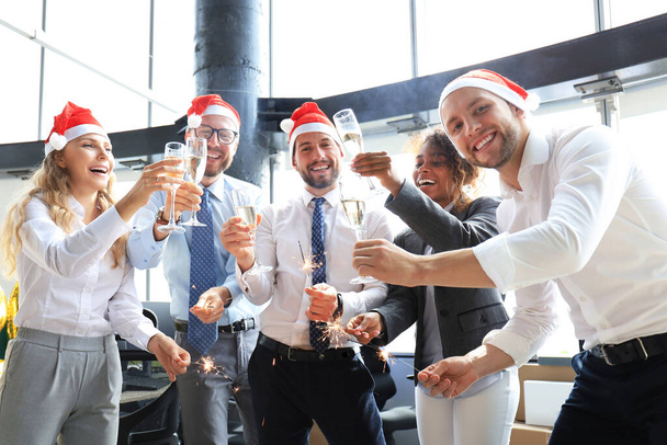 Young business partners are lighting bengal lights and drinking champagne in a modern office. Merry Christmas and Happy New Year - Zdjęcie, obraz