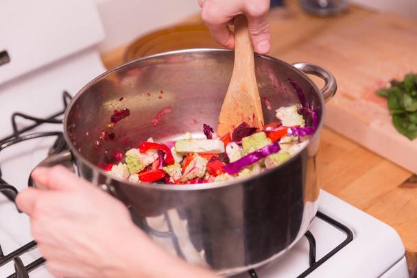 Um cozinheiro mexe legumes em uma panela de aço inoxidável, usando um forno a gás em um ambiente de cozinha em casa. - Foto, Imagem