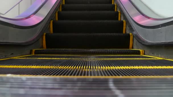低角度の近代的なエスカレーターの階段の視点をループ化した。自動エレベーター機構。紫色の光で照らされた階段の上の黄色の線。未来的な空の機械階段がまっすぐ動く - 映像、動画