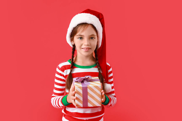 Cute little girl in Santa Claus hat and with Christmas gift on color background - Фото, изображение