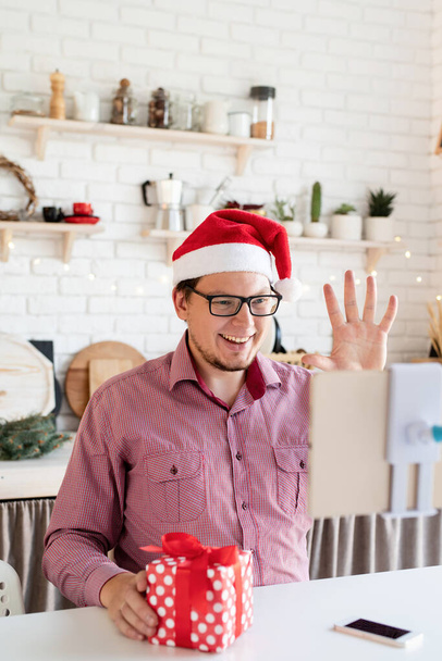 Christmas online greetings. Happy young man in santa hat greeting his friends in video chat or call on tablet sitting in his kitchen - Фото, изображение