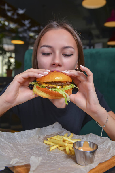 Woman eating burger and fries smiling. Beautiful caucasian female model eating a hamburger with hands over blurred restaurant background. Still life, eating out concept. - Foto, imagen