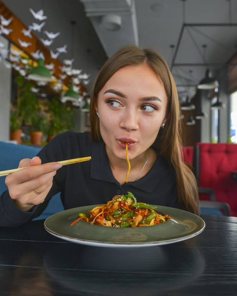Beautiful young woman eating chinese food called Wok with chopsticks. Wok with meat and fried asparagus in a plate. Still life, eating out in restaurant concept. - Foto, afbeelding