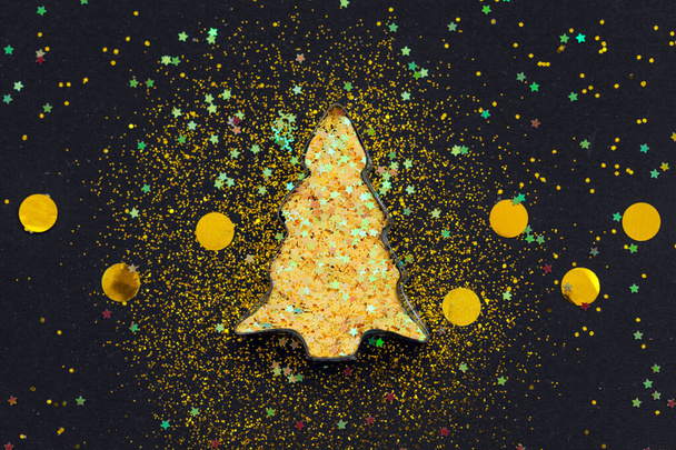Christmas tree shaped cookie cutter with golden sparkles star confetti on black background. Flat lay style. - Fotografie, Obrázek