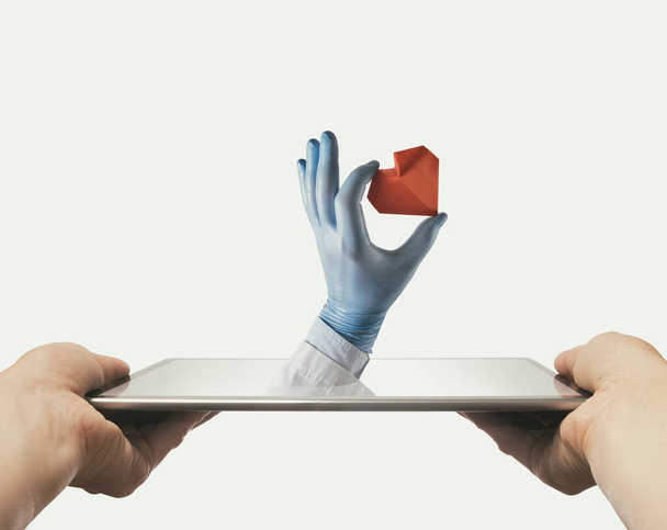 Die Hand des Arztes mit rotem Papierherz ragt aus einem digitalen Tablet-Bildschirm.  - Foto, Bild