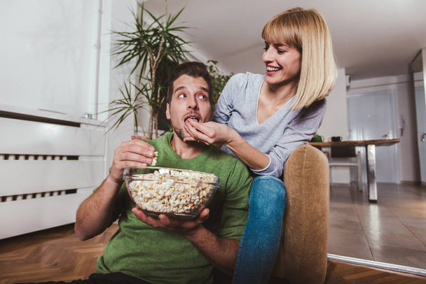 Fiatal pár, akik otthon ülnek a kanapén, tévéznek, boldogan mosolyognak, popcornt esznek, és élvezik az estét.. - Fotó, kép