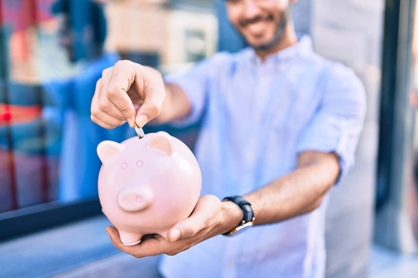 Nuori latino mies hymyilee onnellinen säästää setelin sisällä säästöpossu kaupungin. - Valokuva, kuva