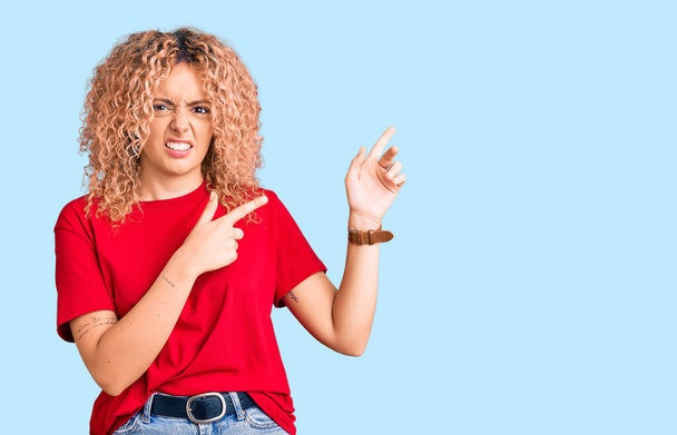 Young blonde woman with curly hair wearing casual red tshirt pointing aside worried and nervous with both hands, concerned and surprised expression  - Fotografie, Obrázek