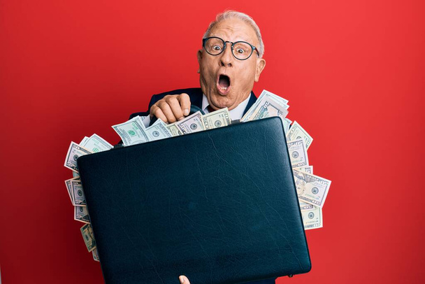 Hombre caucásico mayor sosteniendo maletín lleno de dólares asustado y sorprendido con sorpresa y expresión asombrada, miedo y cara excitada.  - Foto, Imagen