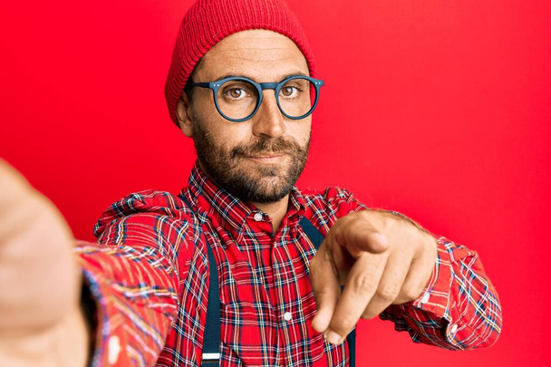 Hombre hipster guapo con barba tomando una foto selfie con el teléfono inteligente apuntando con el dedo a la cámara y a usted, gesto de confianza mirando en serio  - Foto, Imagen