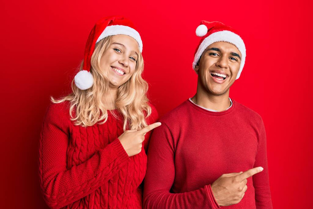 Junges gemischtrassiges Paar mit Weihnachtsmütze, lächelnd und mit zwei Händen und Fingern zur Seite in die Kamera blickend.  - Foto, Bild