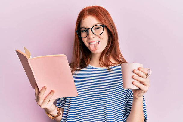 Menina cabeça vermelha jovem lendo um livro e beber uma xícara de café colando a língua para fora feliz com expressão engraçada.  - Foto, Imagem