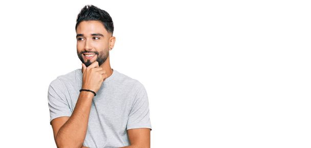 Young man with beard wearing casual grey tshirt with hand on chin thinking about question, pensive expression. smiling with thoughtful face. doubt concept.  - Photo, Image