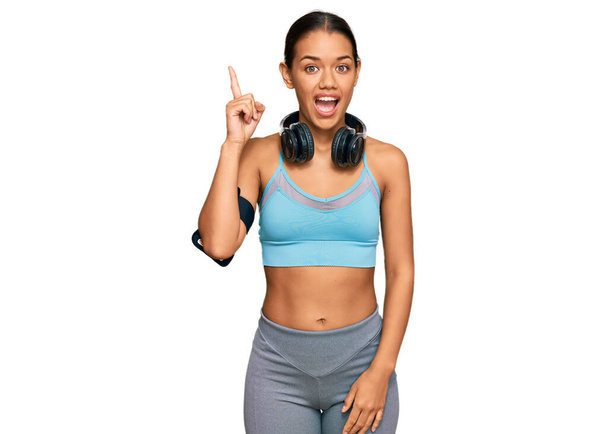Beautiful hispanic woman wearing sportswear and headphones pointing finger up with successful idea. exited and happy. number one.  - Photo, Image