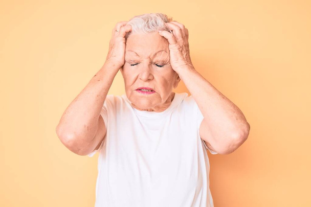 Senior beautiful woman with blue eyes and grey hair wearing classic white tshirt over yellow background suffering from headache desperate and stressed because pain and migraine. hands on head.  - 写真・画像