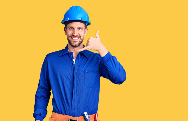 Young handsome man wearing worker uniform and hardhat smiling doing phone gesture with hand and fingers like talking on the telephone. communicating concepts.  - Photo, Image