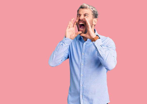 Νέος όμορφος ξανθός άντρας φοράει κομψό πουκάμισο φωνάζει θυμωμένος δυνατά με τα χέρια πάνω από το στόμα  - Φωτογραφία, εικόνα